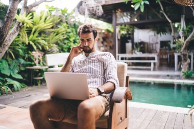 Çizgili gömlekli sakallı genç erkek havuz kenarındaki terasta dinlenirken netbook ekranına bakıyor.