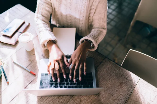 Top view of crop anonymous black female freelancer typing on laptop while sitting at wooden table and working on business project in modern cafe