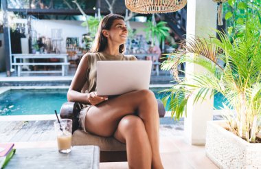 Keyifli etnik kadın rahat koltukta dinleniyor ve Bali 'deki güneşli terasta dizüstü bilgisayarını incelerken başka tarafa bakıyor.