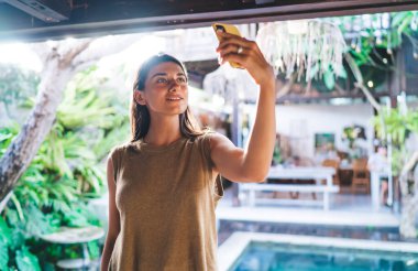 Gündelik giysiler içinde genç bir etnik kadın arka tarafta güneş ışığında yüzme havuzunun yanında kendi portresini çekiyor.