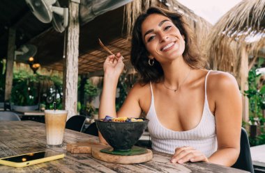 Kolları bükülmüş, kahvaltı kâsesiyle masada otururken kameraya bakan pozitif etnik kadın. 