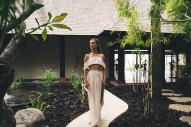 Çıplak ayaklı, şehvetli, beyaz elbiseli bir kadın yolda dikiliyor ve yeşil bahçede yürürken başka tarafa bakıyor.