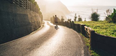 Tanımlanamayan, sıradan kıyafetli, deniz ve dağların yakınındaki boş asfalt yolda yürüyen ve manzaraya hayran olan yalnız bir adamın arka görüntüsü.