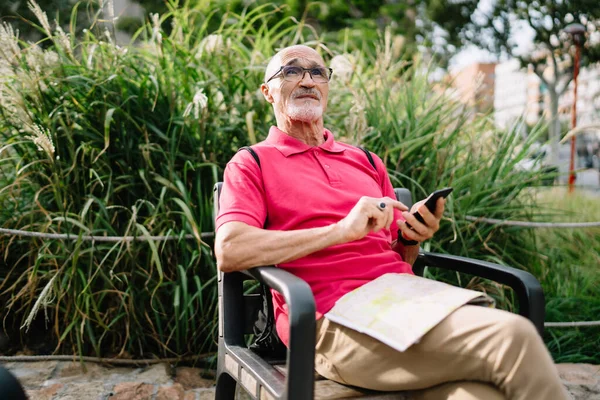 Senior bearded man in casual clothes and glasses sitting on bench near green plants and using smartphone and looking away