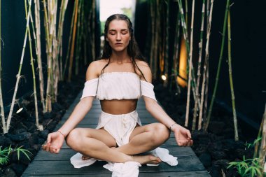 Bahçede meditasyon yaparken Padmasana pozisyonunda bacak bacak üstüne atmış beyaz elbiseli kadın.