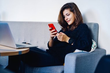 Modayı takip eden, gülümseyen serbest çalışan kadın, akıllı telefondan mesaj okuyor ve masadaki rahat koltukta dizüstü bilgisayarla oturuyor.