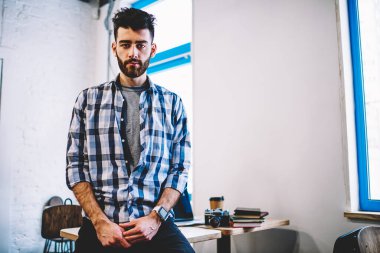 Şık, rahat gömlekler giymiş, fotoğraf makinesine bakan ciddi bir gencin portresi. Kendi tasarım stüdyosunun içinde, masaüstünde duran yaratıcı hipster fotoğrafçı.