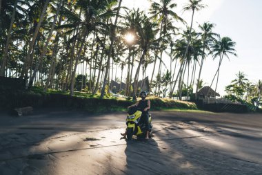 Full length of unrecognizable male biker wearing protective helmet driving motorcycle along empty sandy beach near palm trees on sunny day