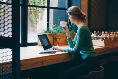 Kafe projesinde çalışırken tahta masada dizüstü bilgisayar ve bir fincan kahveyle oturan serbest çalışan serbest çalışanın yan görüntüsü.