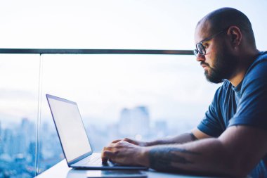 Gözlüklü, panoramik pencerenin yanındaki masada çalışırken dizüstü bilgisayar kullanan odaklanmış bir erkeğin yan görüntüsü.