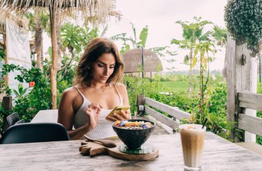 Yazın kafeterya verandasında lezzetli yemekler ve bir bardak kahveyle cep telefonunu izleyen genç etnik bayan.