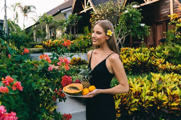 トロピカルガーデンの赤い花の近くに立っている間に新鮮な果物を保持黒のドレスを身に着けている笑顔の女性の側面図 — ストック写真