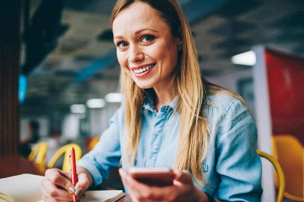 Crop of young female with toothy smile looking at camera sitting at table with smartphone taking notes in planner while having break