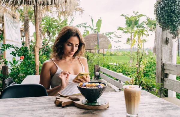 Yazın kafeterya verandasında lezzetli yemekler ve bir bardak kahveyle cep telefonunu izleyen genç etnik bayan.
