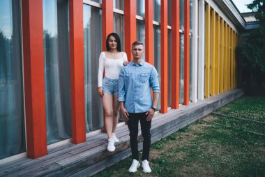 Modern binanın yanında dikilen ve birlikte vakit geçirirken kameraya bakan çok ırklı, ciddi bir çift.