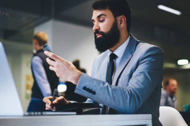 Resmi takım elbiseli, konsantre sakallı iş adamlarından dizüstü bilgisayarlı, defterli ve modern ofisteki akıllı telefonları tarayan.