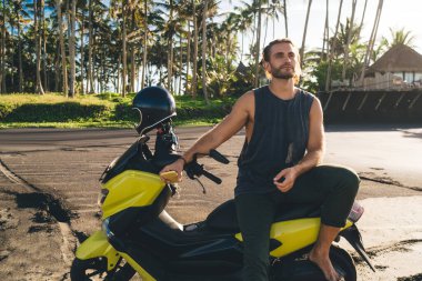 Positive male hipster with smartphone sitting on motorcycle and looking away while resting alone in countryside and enjoying rural trip