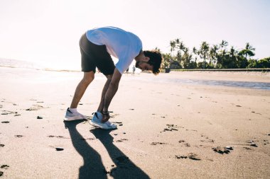Eğitimler için rahat giysiler içinde sakallı genç bir adamın güneş ışığında kumlu sahilde durup ayakkabı bağcıklarını bağlamasının yan görüntüsü.