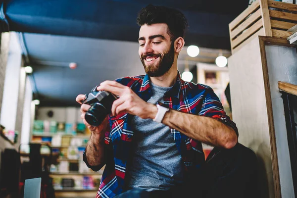 Cheerful bearded young man laughing during viewing funny photos sitting in own design studio.Positive hipster guy working photographer holding vintage camera in hand and watching video on display