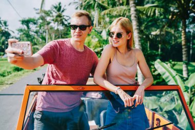 Genç bayan ve erkek, güneşli bir yaz gününde tropikal bir ülkede seyahat ederken ve kendi fotoğrafını çekerken güneş gözlüğü takarken açık arabada duruyorlar.