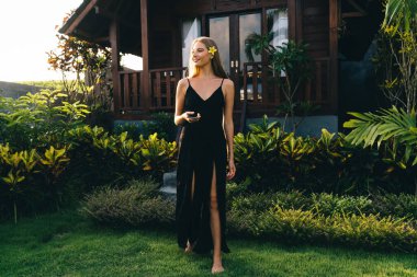 Güneşli bir günde bahçede egzotik bitkilerle dikilirken siyah elbiseli, gülümseyen bir kadın cep telefonu kullanıyor.