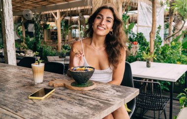 Dışarıdaki kafede kahvaltı kasesi ve bir bardak kahveyle masaya oturmuş kameraya bakan pozitif etnik bir kadın.