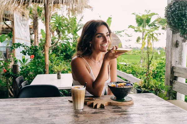 Happy smiling female in summer wear recording voice message on modern cellphone while having delicious meal in tropical outdoor lounge