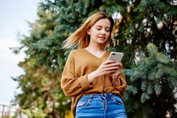 From below of focused young female in jeans and cardigan text messaging with friend on smartphone while standing near coniferous trees in park
