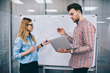 Modern ofiste, beyaz tahtaya yakın duran ve dizüstü bilgisayar ve çizelge kullanarak günlük giysiler giyerek proje üzerinde çalışan genç iş arkadaşlarının yan görüntüsü