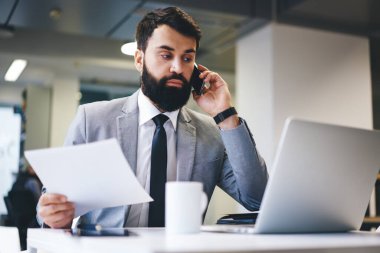 Resmi takım elbiseli sakallı konsantre işadamı dizüstü bilgisayarlı ve belgeli masada oturuyor ve hafif modern çalışma alanında akıllı telefondan arıyor.