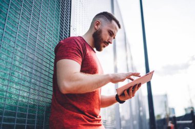 Sokakta metal çitlere yaslanırken kırmızı gömlekli, sakallı, sörf tabletli düşünceli bir erkeğin yan görüntüsü.