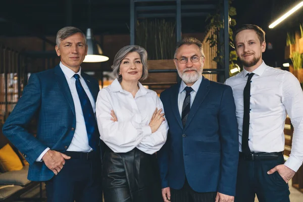 Confident mature woman and men in suits standing together in modern office workplace with modern interior and looking at camera