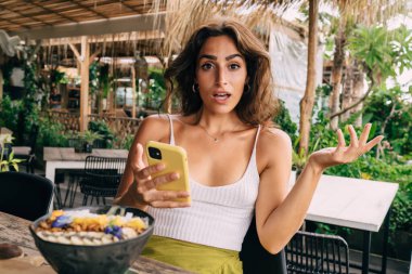 Bilmece gibi giyinmiş genç bir kadın cep telefonu kullanıyor ve tropik bir restoranda dinlenirken hayret verici bir şekilde kameraya bakıyor.