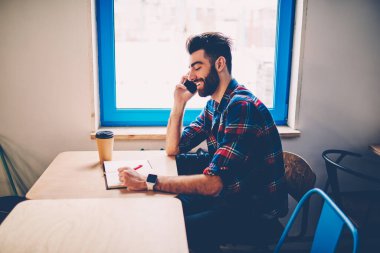 Sıradan giyinmiş sakallı bir gencin telefon görüşmeleri sırasında telefon görüşmeleri ve kahkahalar atarak not almasının yan görüntüsü. 