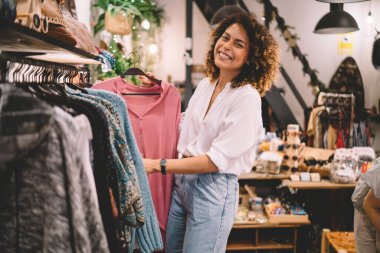 Moda kıyafetleri içinde neşeli bir kadın şık kıyafetler alıyor ve askı takarken kameraya gülümsüyor.