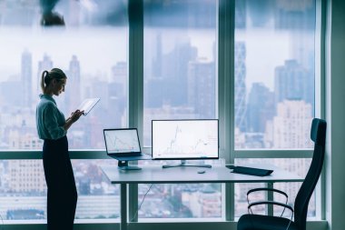 Modern ofisin penceresinin yanında dizüstü bilgisayarla gezen şık günlük kıyafet tabletindeki memnun yönetici kadının yan görüntüsü