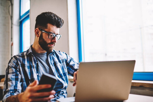 Serious hipster guy sitting at university campus spending time with modern laptop computer and waiting friend indoors