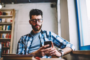 Üniversitede oturan gözlüklü genç adam cep telefonuna uygulama yüklüyor.