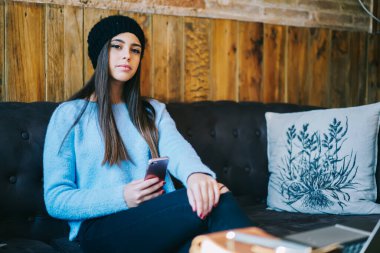 Kafkas kadın blogcunun portresi modern cep telefonu cihazını elinde tutuyor ve boş zamanlarında rahat koltukta kameraya bakıyor.
