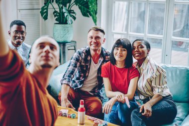 Şık casual giyinen mutlu çok kültürlü hipster rahat kanepede oturan ve dostça toplantı sırasında fotoğraf yaparken gülümseyen giyiyor. Birlikte vakit olumlu gençlerin