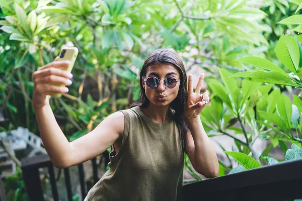 エキゾチックな植物の近くの熱帯リゾートでアヒルの顔と平和のサインを作りながら スマートフォンで自撮りサングラスの正の民族の女性 — ストック写真