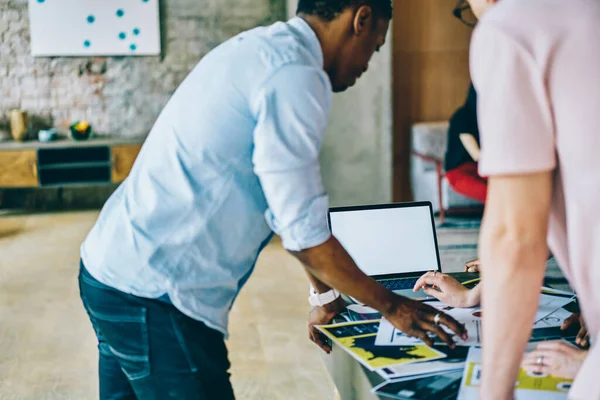 Crop of African American male programmer in casual formal wear helping colleagues with mistake on empty screen laptop at stylish office