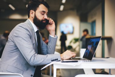 Resmi takım elbiseli, sakallı, netbook 'la masada oturan ve ofiste çalışırken cep telefonuyla konuşan çalışanın yan görüntüsü.
