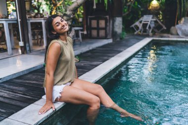 Eğimli kafası ve dişlek gülüşü olan neşeli etnik kadın yüzme havuzunun kenarında oturmuş, bacakları suda kameraya bakıyor.