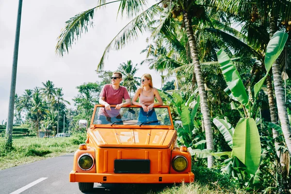 Genç bir erkek ve kadın turuncu renkli bir arabada güneşli bir yaz gününde seyahat ederken ve başka tarafa bakarken güneş gözlüğü takarken.
