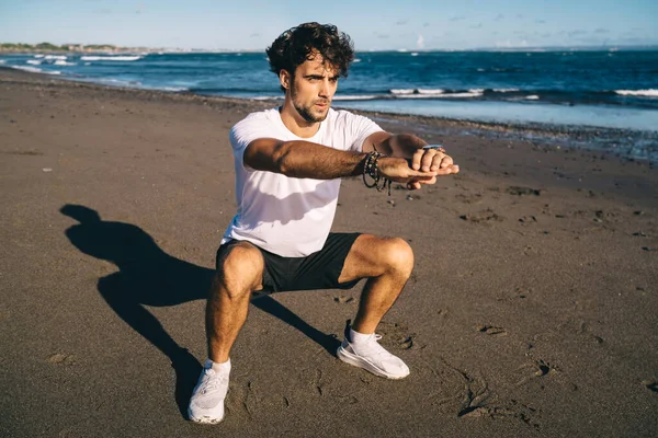 Full body of bearded male runner wearing sportswear and sneakers doing squats before jogging on seashore on summer day and looking away