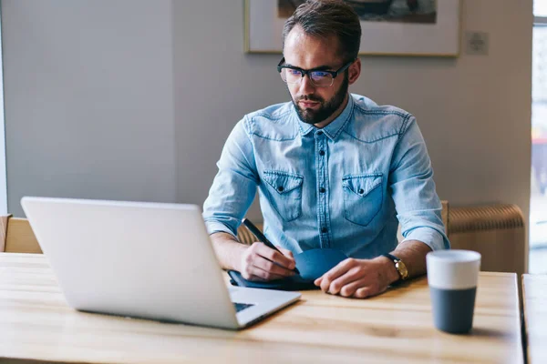 Skoncentrowany Mężczyzna Freelancer Dżinsowej Koszuli Patrząc Laptopa Robi Notatki Siedząc — Zdjęcie stockowe