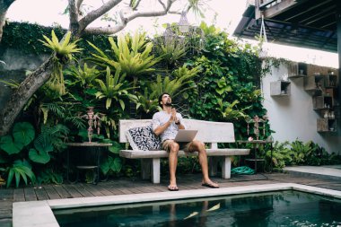 Uzun boylu, gergin bir erkek. Yaz kıyafeti giyiyor. Yüzme havuzunun yanındaki bankta oturuyor.