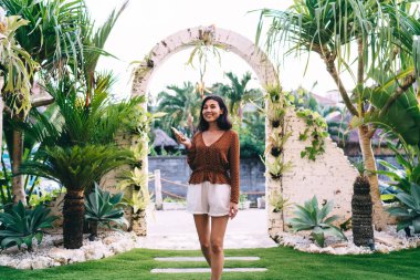 Şık elbiseli mutlu esmer kadın akıllı telefona göz gezdirirken evinin yakınındaki kemerli yolda yürürken kameraya bakıyor.