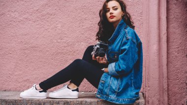 Uzun kıvırcık saçlı, kot ceketli, siyah pantolonlu, taş bankta oturan, akıllı telefonlu ve elinde retro fotoğraf makinesi olan kendine güvenen genç bir kadın.